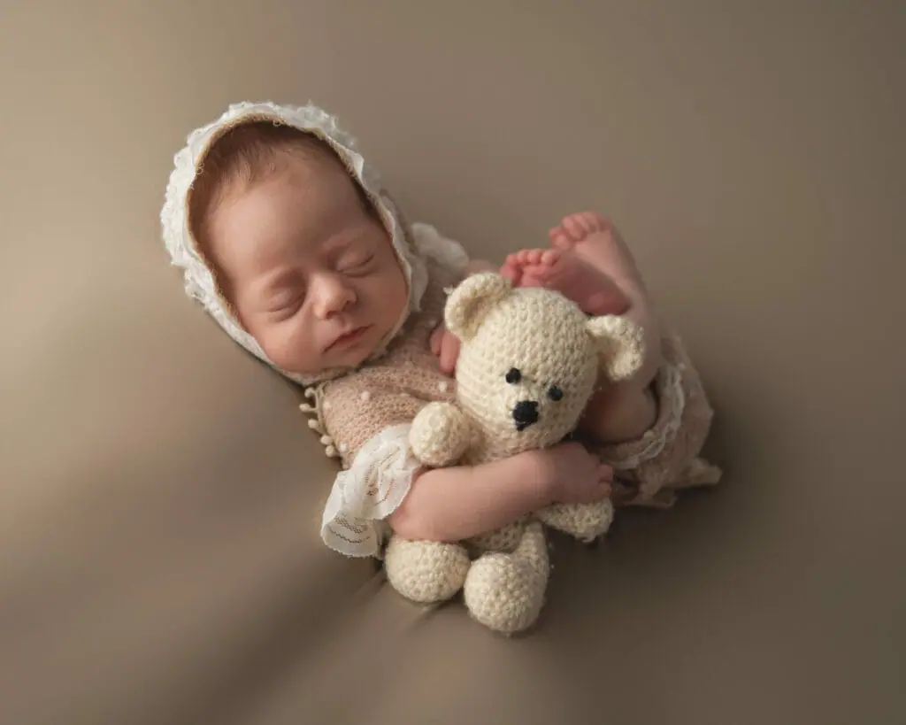 A newborn baby sleeps peacefully while wearing a white knit bonnet and holding a crocheted teddy bear. The baby has eyes closed and is wrapped in a neutral-colored blanket.