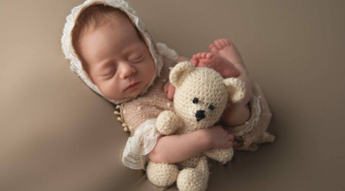 A newborn baby sleeps peacefully while wearing a white knit bonnet and holding a crocheted teddy bear. The baby has eyes closed and is wrapped in a neutral-colored blanket.