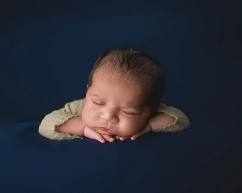 Newborn baby peacefully sleeping with head resting on hands against a dark blue background.