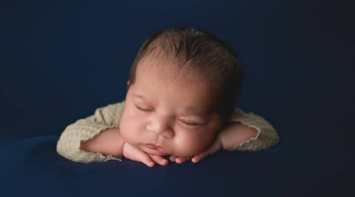 Newborn baby peacefully sleeping with head resting on hands against a dark blue background.