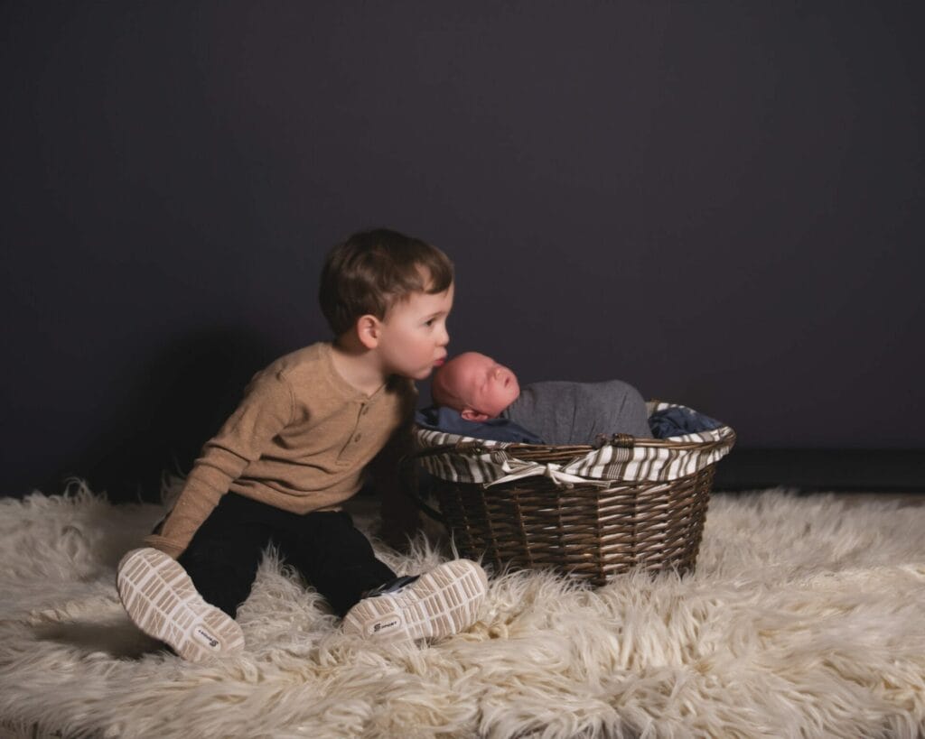 A young child sitting on a white rug leans towards a newborn baby wrapped in a gray blanket, resting in a wicker basket. The background is dark, highlighting the children.