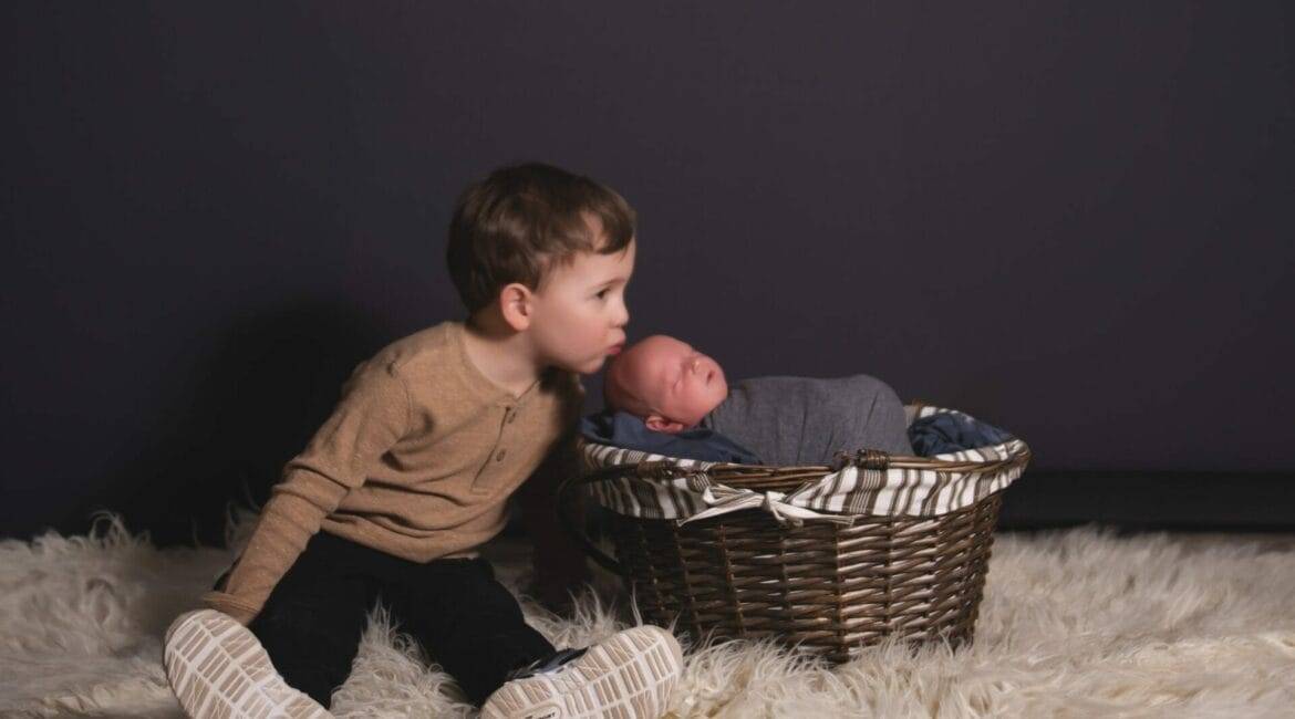 A young child sitting on a white rug leans towards a newborn baby wrapped in a gray blanket, resting in a wicker basket. The background is dark, highlighting the children.