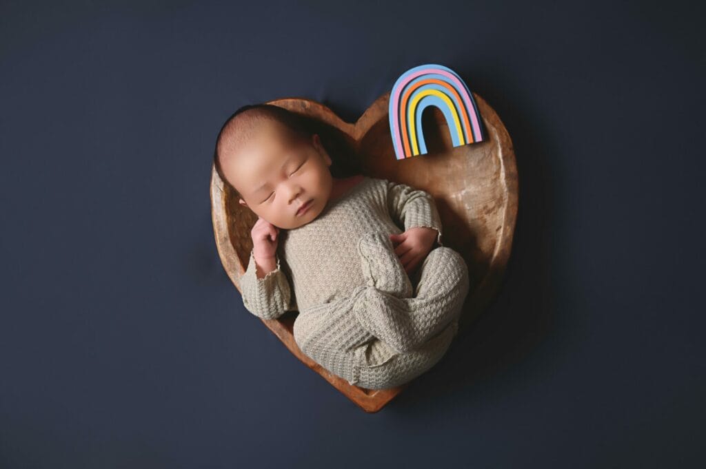 A baby sleeps in a heart-shaped wooden bowl with a small rainbow figure beside them on a dark background.