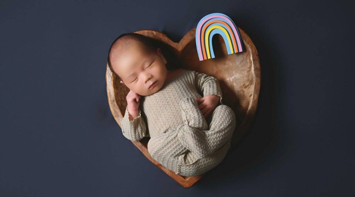 A baby sleeps in a heart-shaped wooden bowl with a small rainbow figure beside them on a dark background.