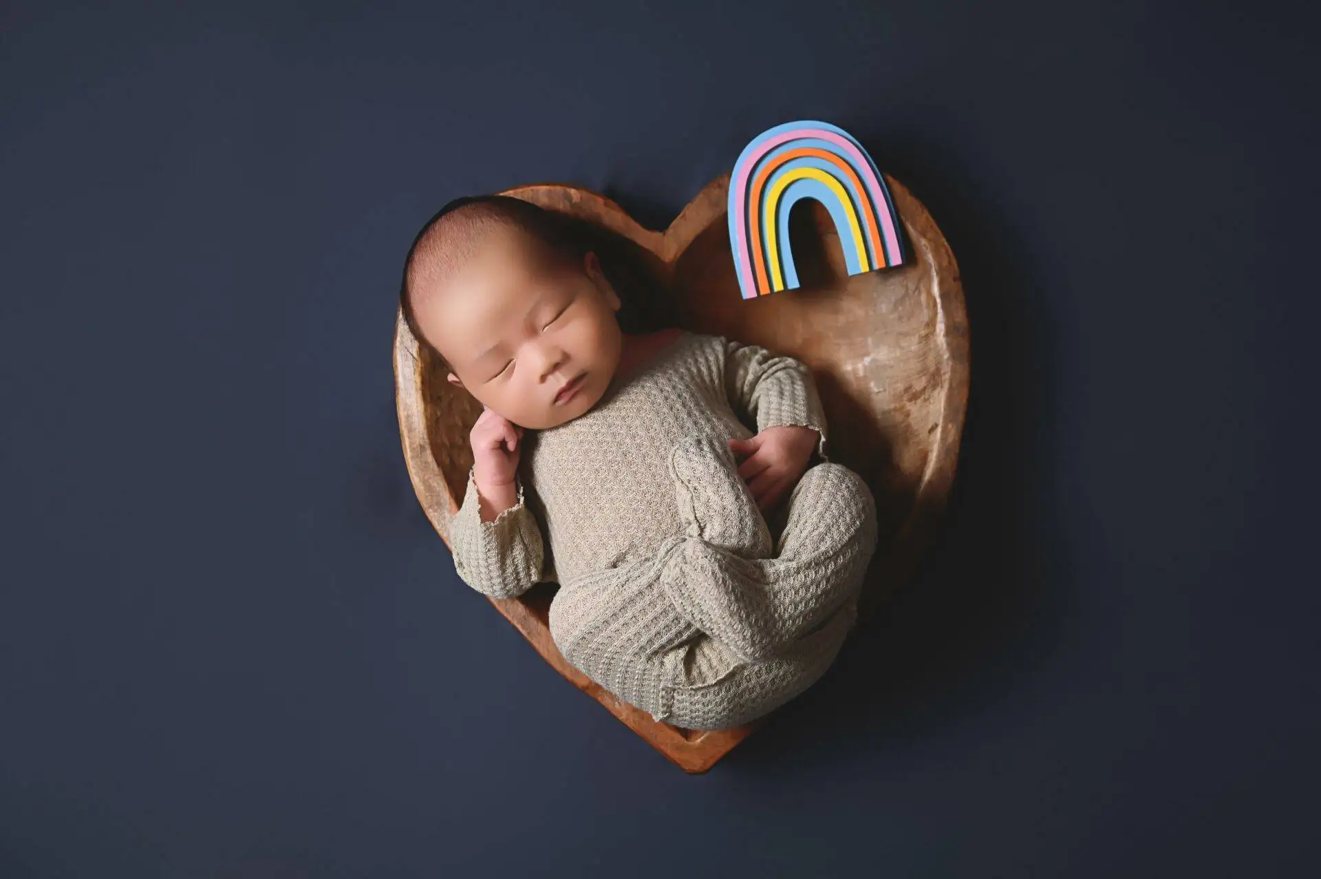 A baby sleeps in a heart-shaped wooden bowl with a small rainbow figure beside them on a dark background.