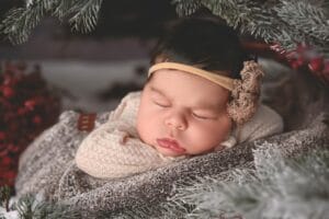 A sleeping baby with a headband is nestled in a cozy, textured blanket surrounded by pine branches.