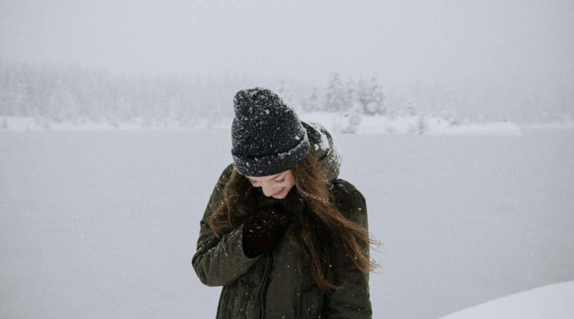 A person in a winter coat and beanie stands in a snowy landscape, looking down and smiling.