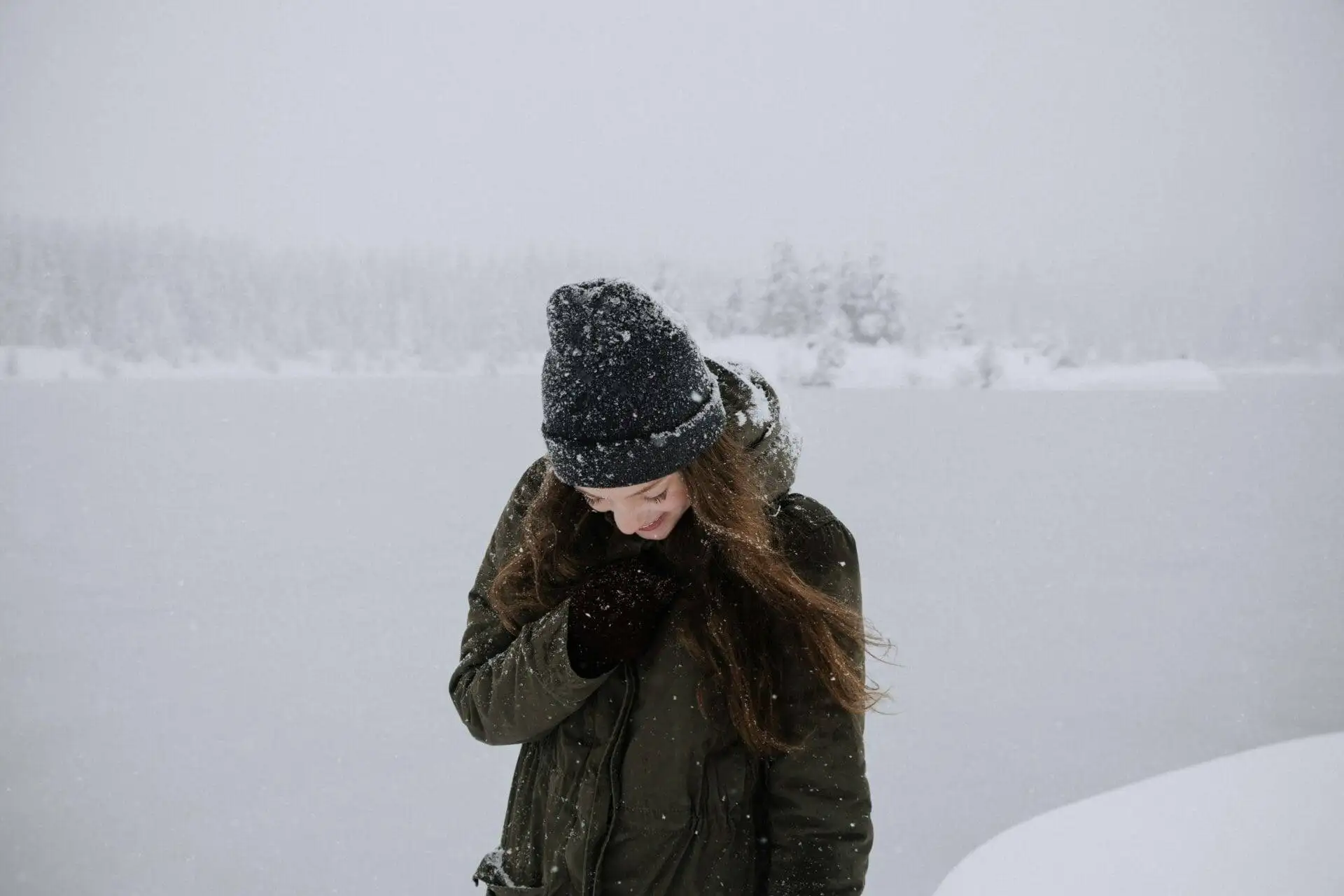 A person in a winter coat and beanie stands in a snowy landscape, looking down and smiling.