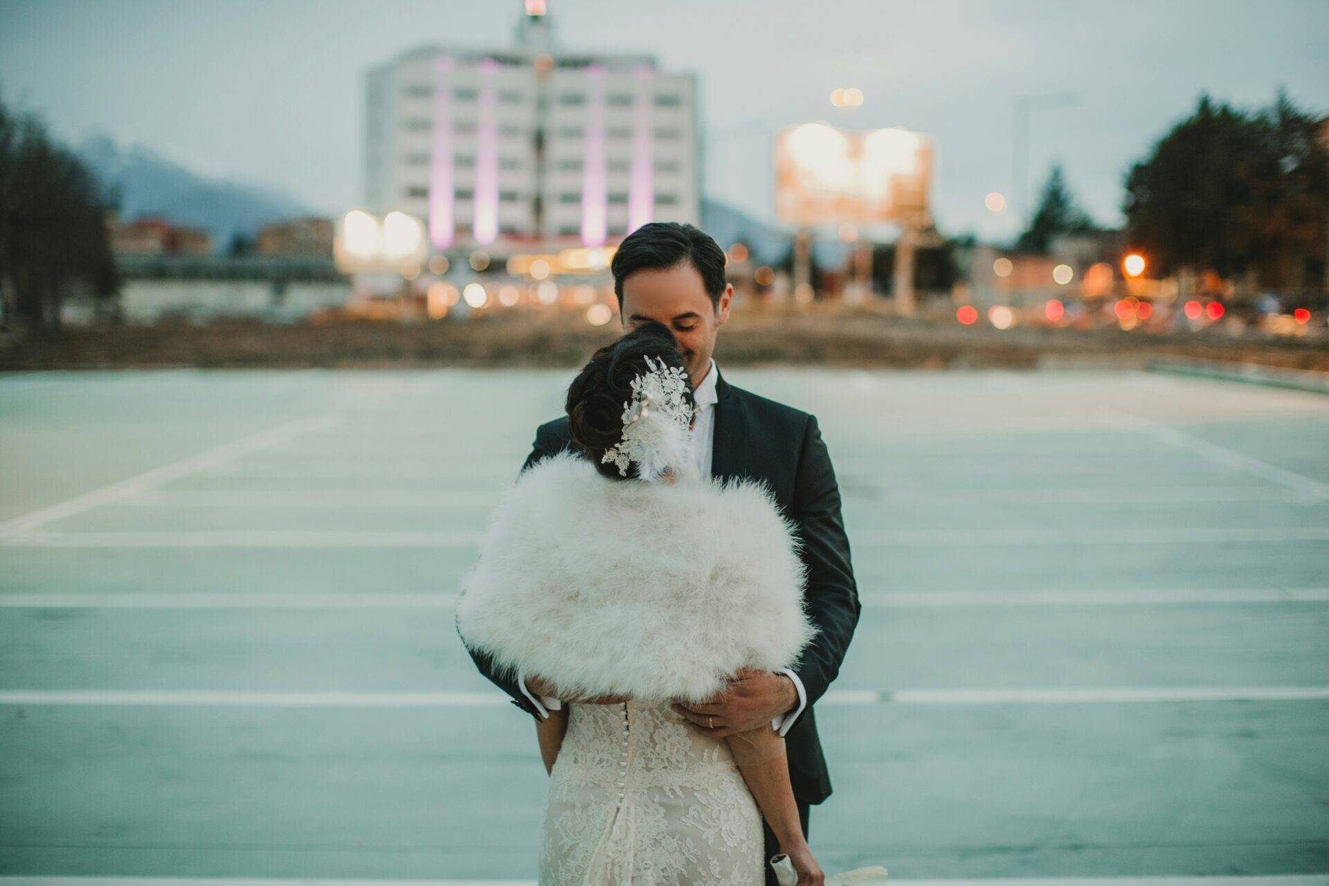 A couple embraces outdoors in formal attire, with the woman wearing a white dress and fur shawl. The background features a blurred building with lights.