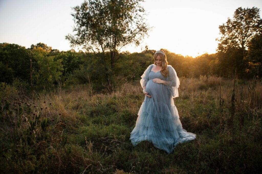 A pregnant woman in a flowing blue dress stands in a grassy field at sunset, surrounded by trees.
