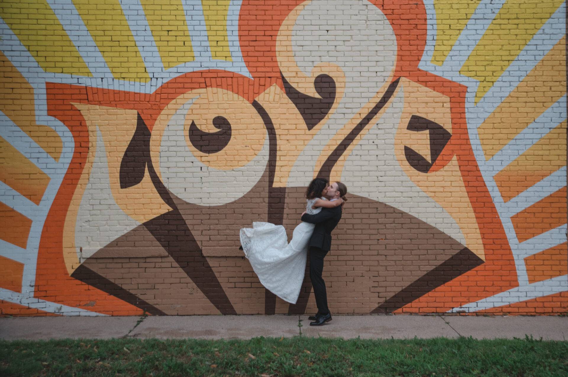 A couple embraces and kisses in front of a colorful mural with the word "love" on a brick wall. The background features orange and yellow rays.