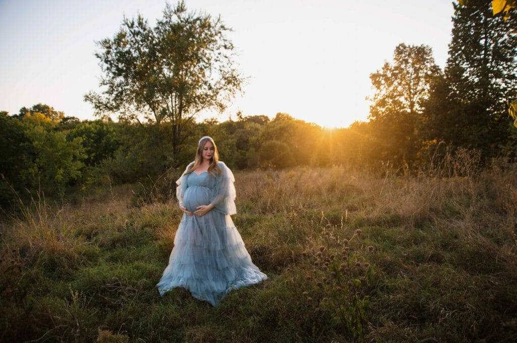 A pregnant woman in a flowing gown stands in a field at sunset, surrounded by trees and tall grass.