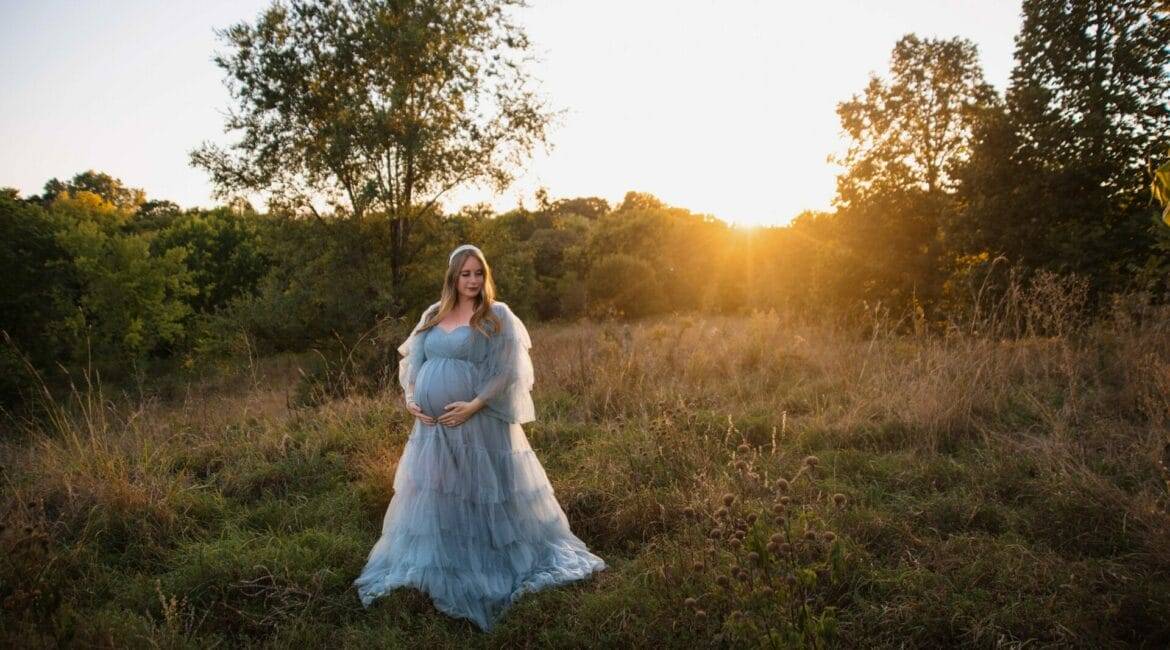 A pregnant woman in a flowing gown stands in a field at sunset, surrounded by trees and tall grass.