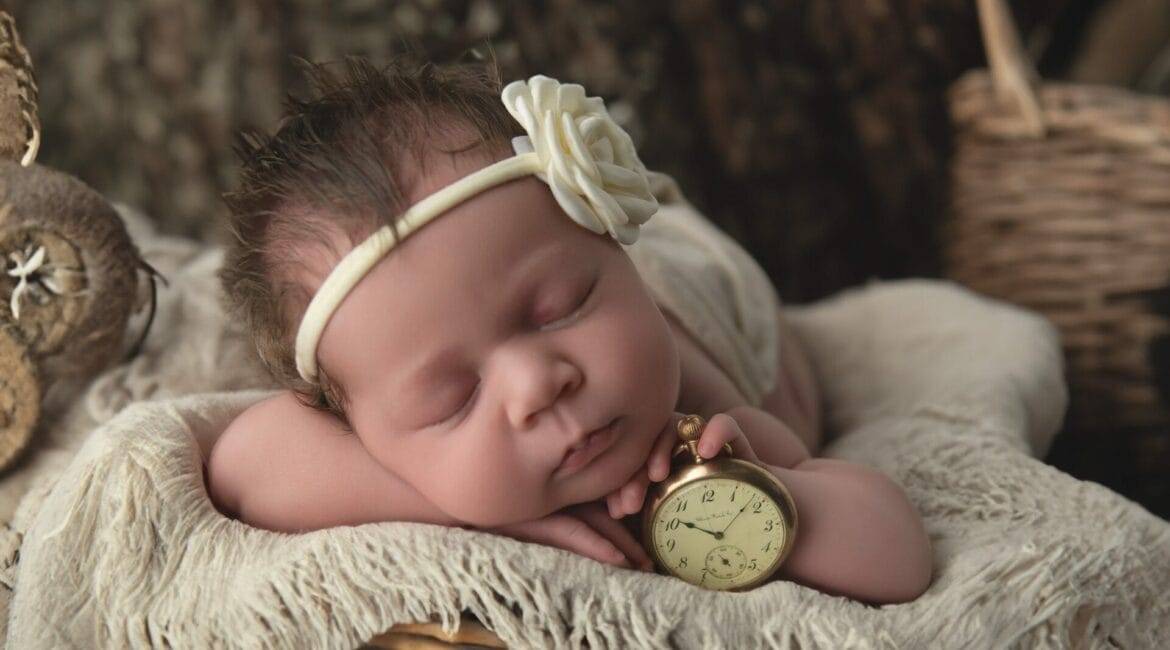 Sleeping baby with a headband and flower, lying on a soft blanket in a wicker basket, holding an antique pocket watch.