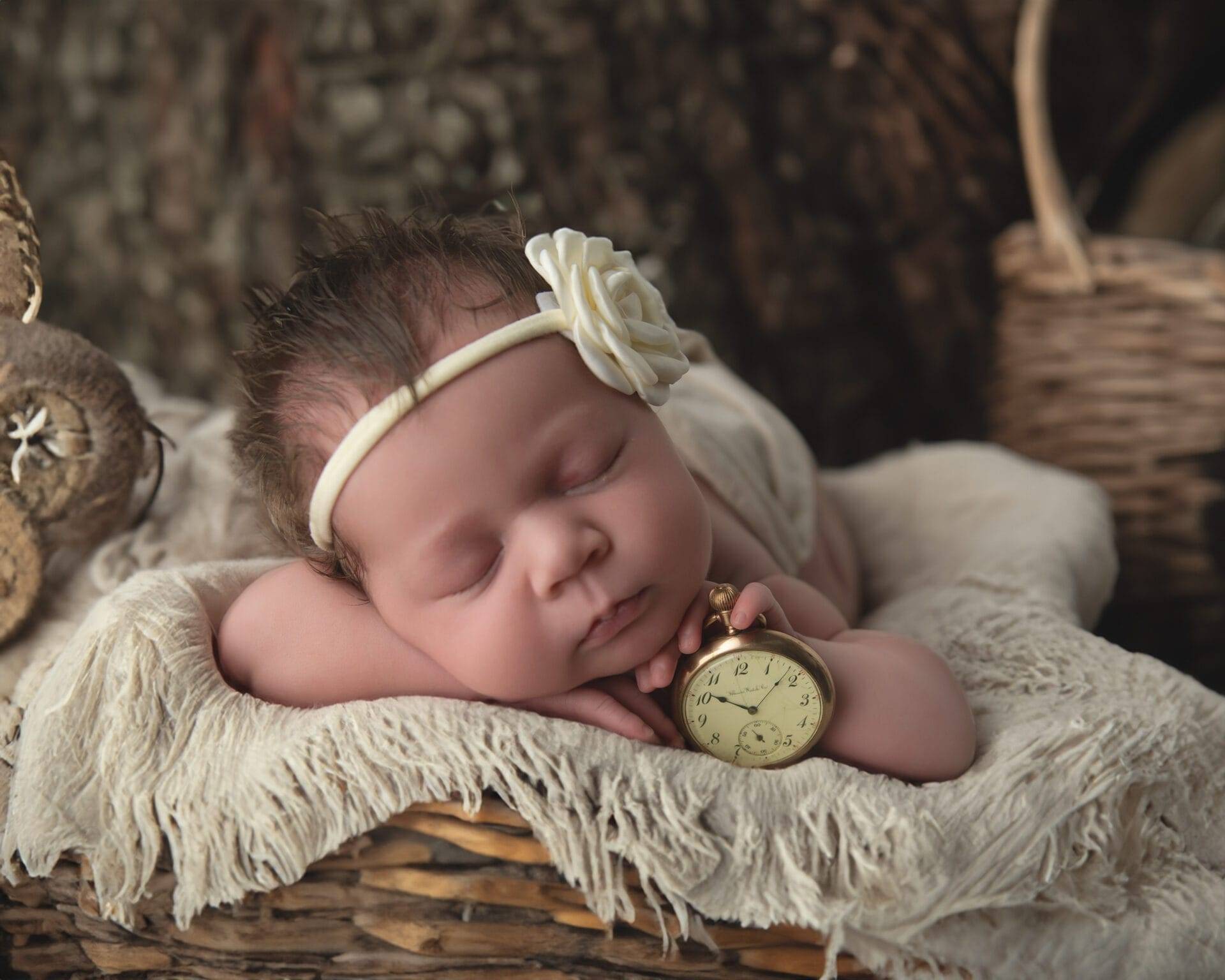 Sleeping baby with a headband and flower, lying on a soft blanket in a wicker basket, holding an antique pocket watch.