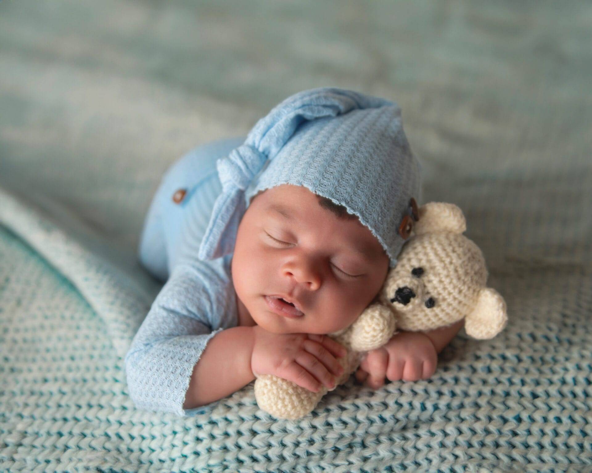 A sleeping baby in blue pajamas and hat, resting on a knitted blanket, cuddles a small crocheted teddy bear.