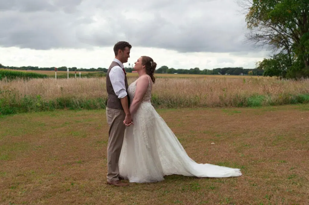 A bride and groom stand holding hands and facing each other in an open field, under a cloudy sky.