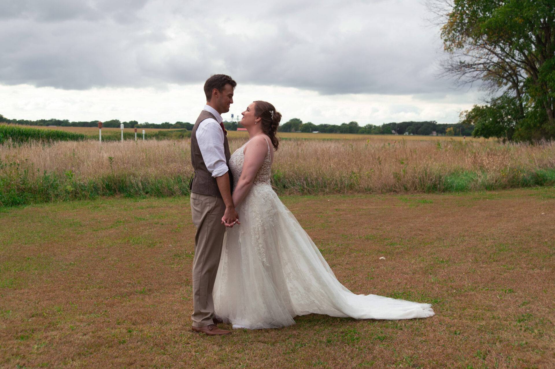 A bride and groom stand holding hands and facing each other in an open field, under a cloudy sky.