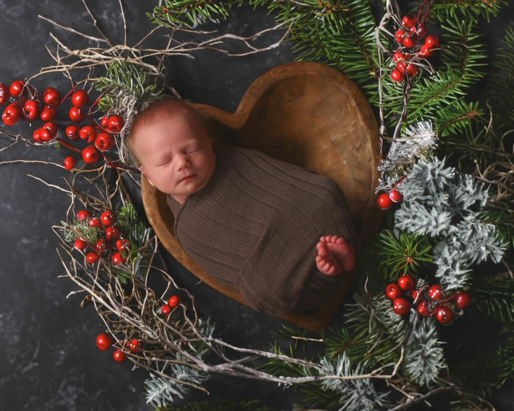 A newborn baby wrapped in brown fabric sleeps in a heart-shaped wooden bowl, surrounded by greenery and red berries on a dark background.