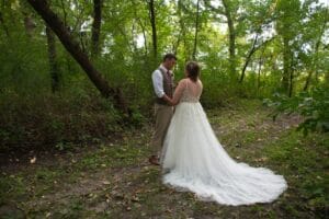 A bride and groom hold hands in a forested area, surrounded by lush green trees. The bride wears a white gown with a long train, and the groom is in a vest and tie.
