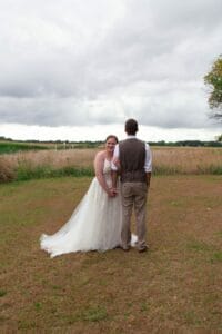 A bride and groom stand in a grassy field, with the bride facing the camera and the groom facing away. The sky is overcast.