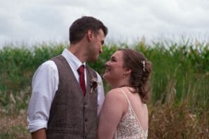 A couple stands outdoors, facing each other. The man wears a vest and tie, and the woman is in a lace dress. They are surrounded by tall grass, with a cloudy sky above.