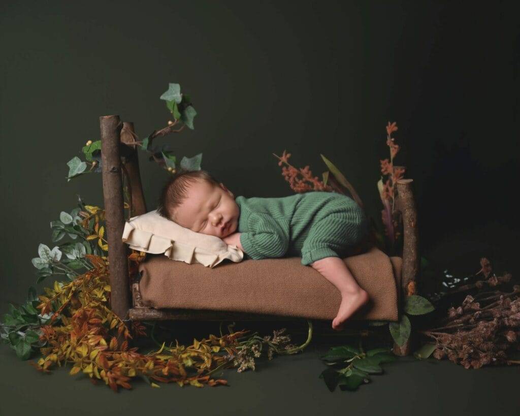 A sleeping baby in a green outfit lies on a small wooden bed surrounded by greenery against a dark background.