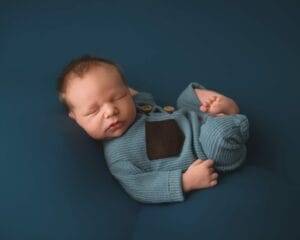 A sleeping baby wearing a blue knitted outfit with a dark patch lies on a dark blue surface.