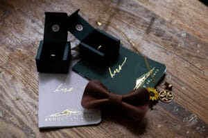 Wooden table with wedding items: ring boxes, vows booklets labeled "hers", a brown bow tie, and gold jewelry including a sunflower pendant.