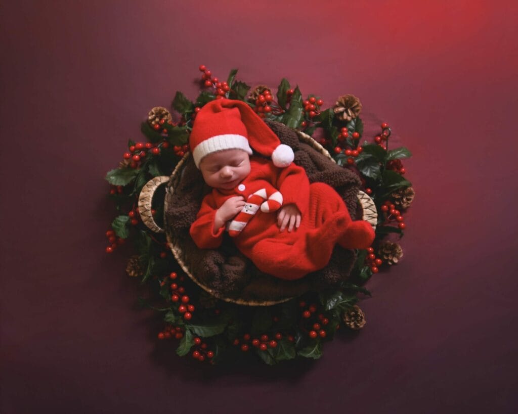 A newborn baby in a red outfit and Santa hat sleeps in a basket surrounded by holly leaves, red berries, and pine cones, set against a red background.