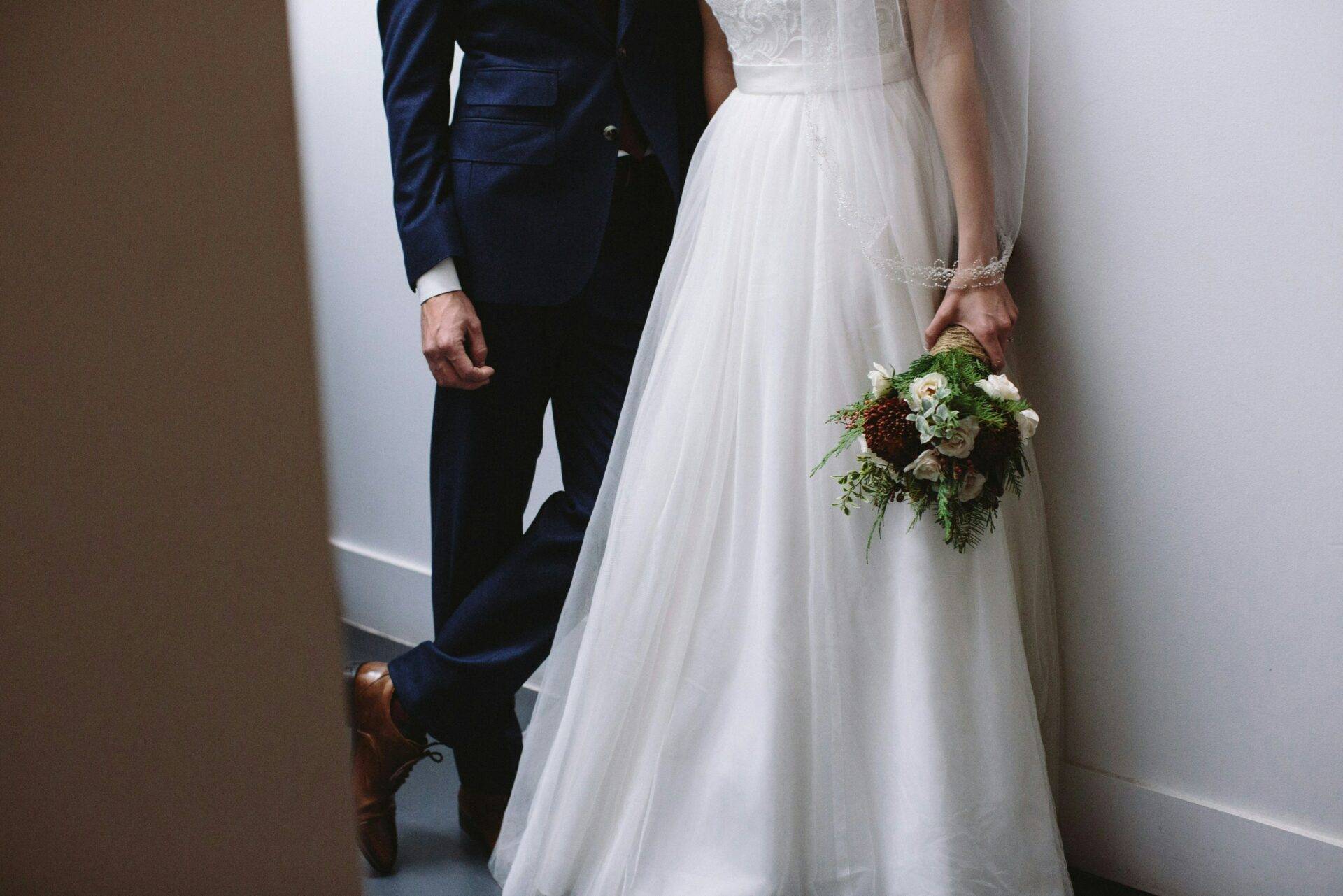 A bride and groom stand closely, leaning against a wall. The bride holds a bouquet of flowers. The groom is in a dark suit and brown shoes.