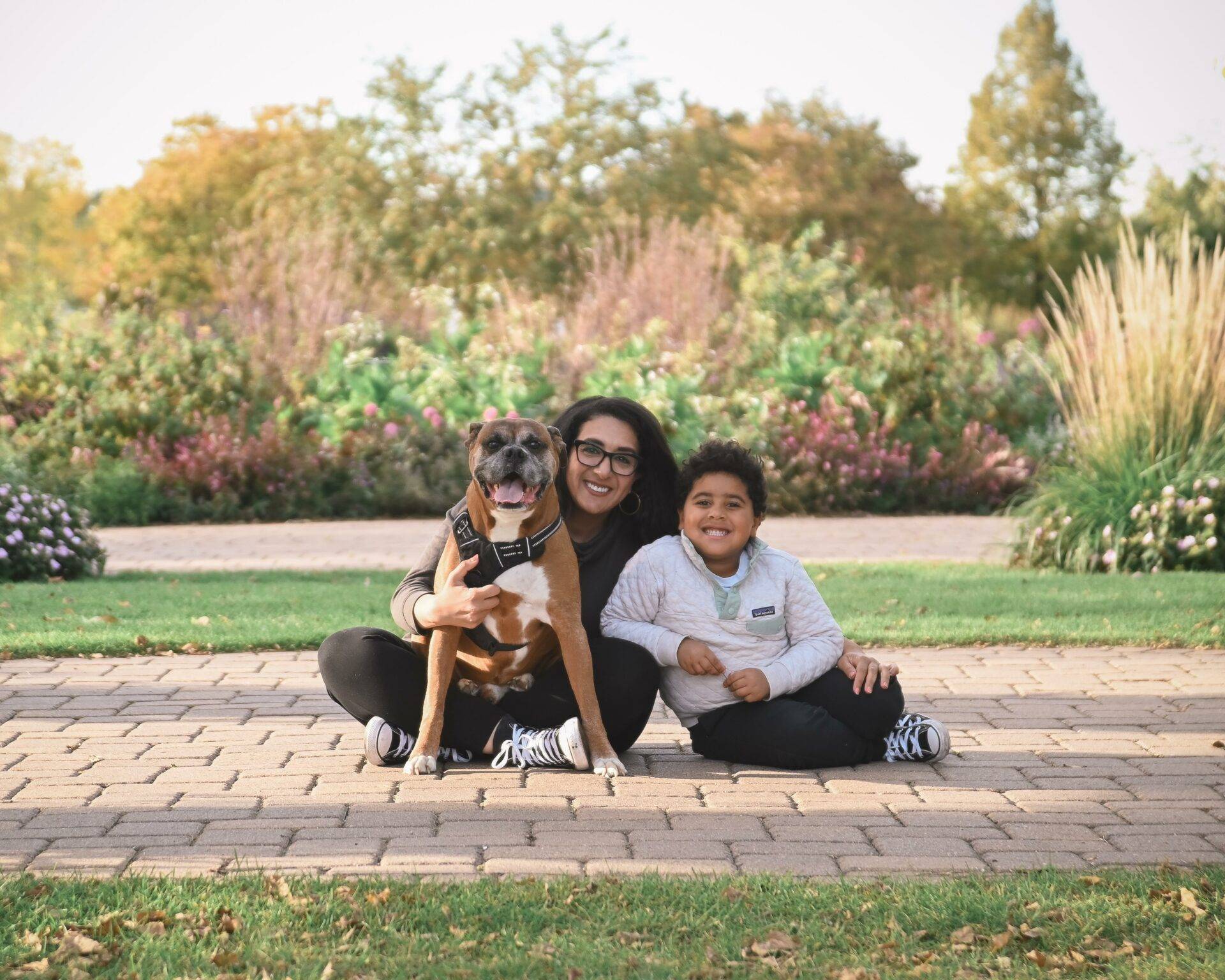 A person, child, and dog sit on a brick path in a park. The child and dog wear matching clothes. Greenery and trees are in the background.