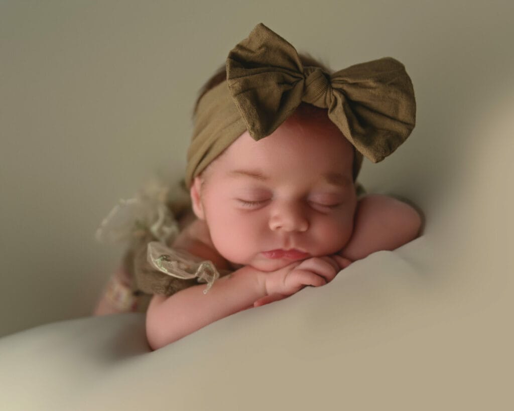 A sleeping baby with a large brown bow headband rests their head on folded arms.