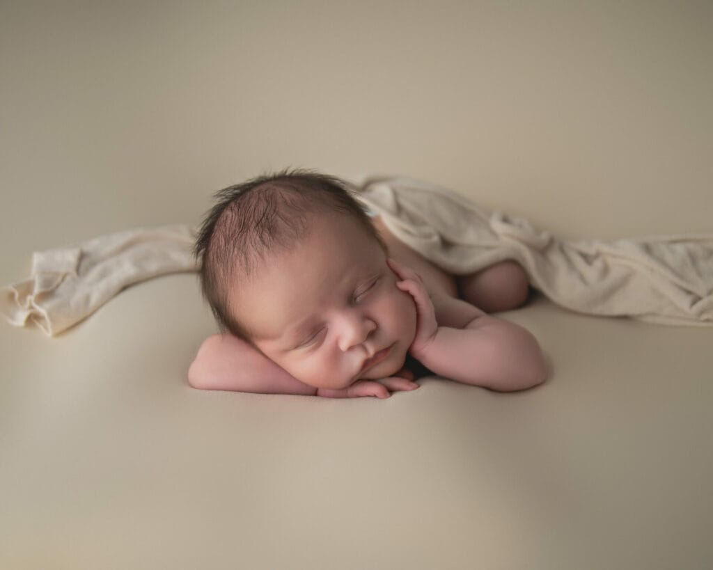 A sleeping baby rests with its head on its hands and a light cloth draped over its back, set against a soft beige background.