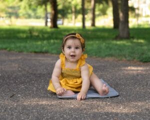 A baby in a yellow dress sits on a gray mat on a paved path in the park, surrounded by green trees, creating an enchanting scene of children photography.