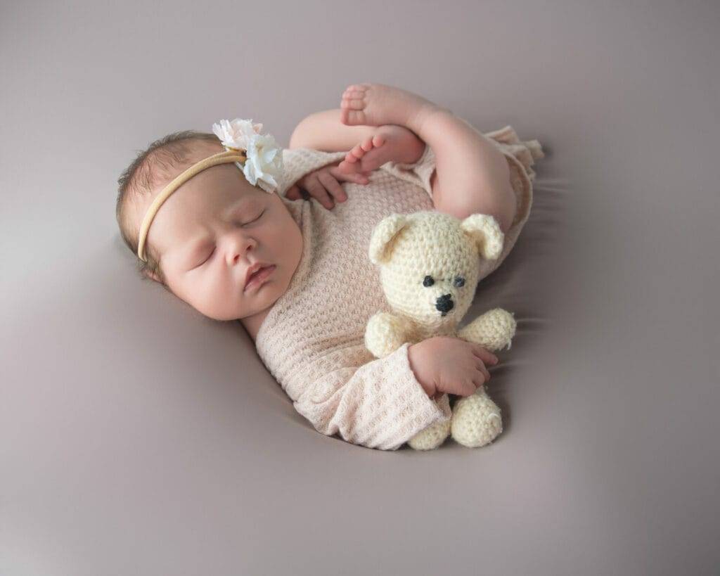 Newborn baby sleeping peacefully, wearing a light outfit with a headband, holding a small knitted teddy bear, against a neutral background.