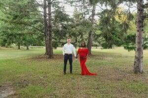 A couple stands hand in hand in a grassy park surrounded by trees. The man wears a white shirt and dark pants, while the woman is in a red dress.