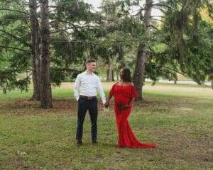 A couple stands holding hands in a grassy area surrounded by trees. The woman is wearing a red dress, and the man is in a white shirt and dark pants.