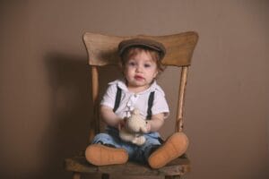 In a charming example of children photography, a toddler in a brown cap and suspenders sits on a wooden chair, clutching a knit toy against a plain backdrop.