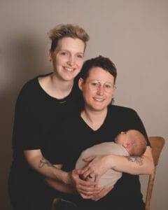 Two smiling individuals with short hair sit together. One holds a sleeping baby wrapped in a beige blanket. Both wear black shirts and have visible tattoos on their arms.