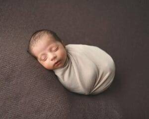 A sleeping newborn baby swaddled in a light beige blanket lies on a dark brown textured surface.