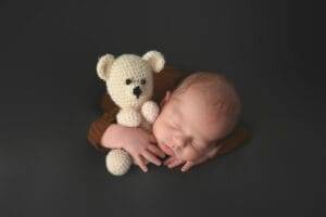 A sleeping newborn baby cradles a small, knitted bear on a dark surface.