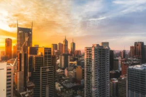 City skyline at sunset, featuring tall buildings with the sun casting a warm glow on the architecture and scattered clouds in the sky.