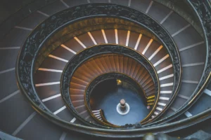 Spiral staircase with ornate railing, viewed from above, showing concentric circular patterns and steps leading down to a central column.