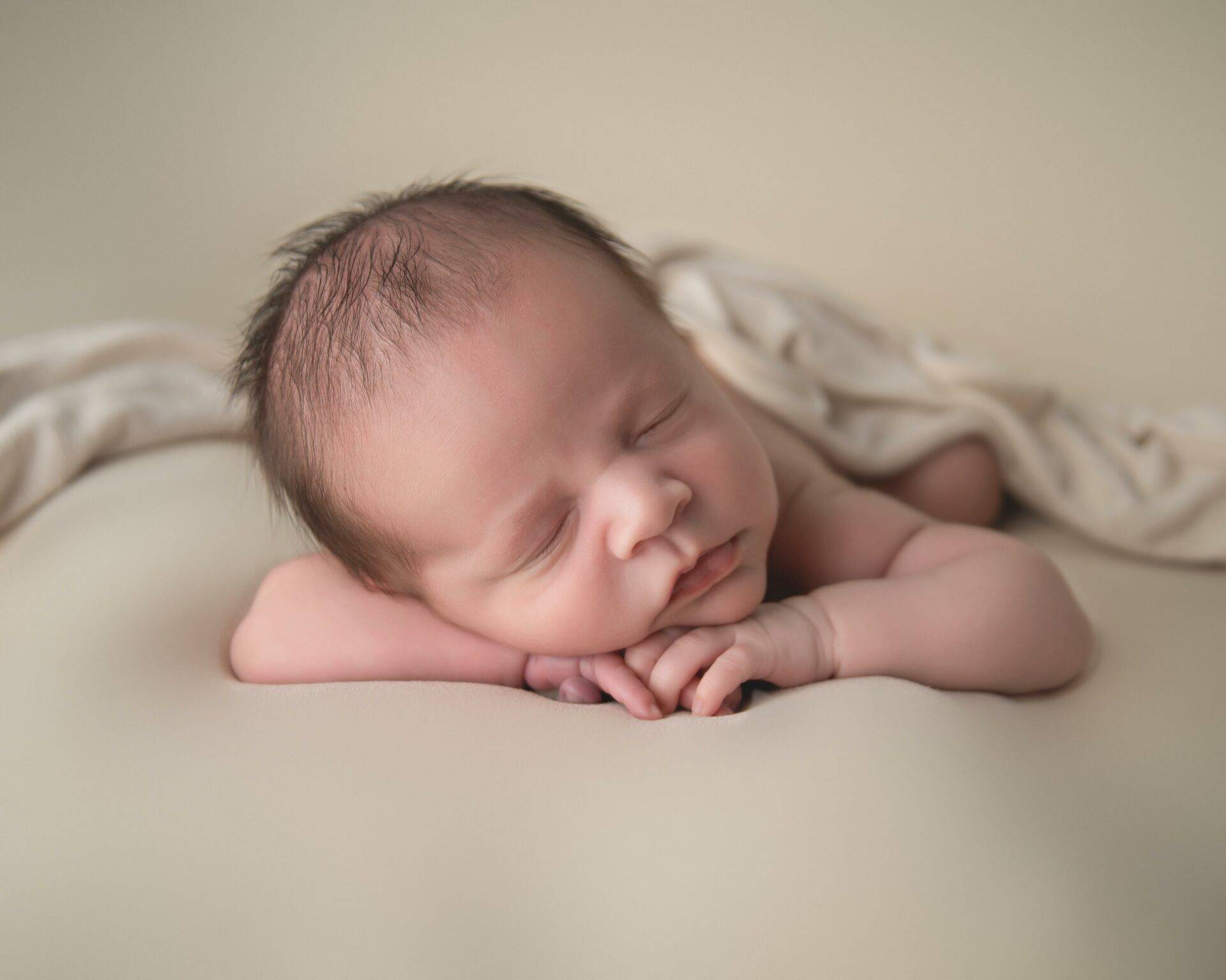 Newborn baby sleeping peacefully on a soft beige surface with a light blanket partially covering their back.