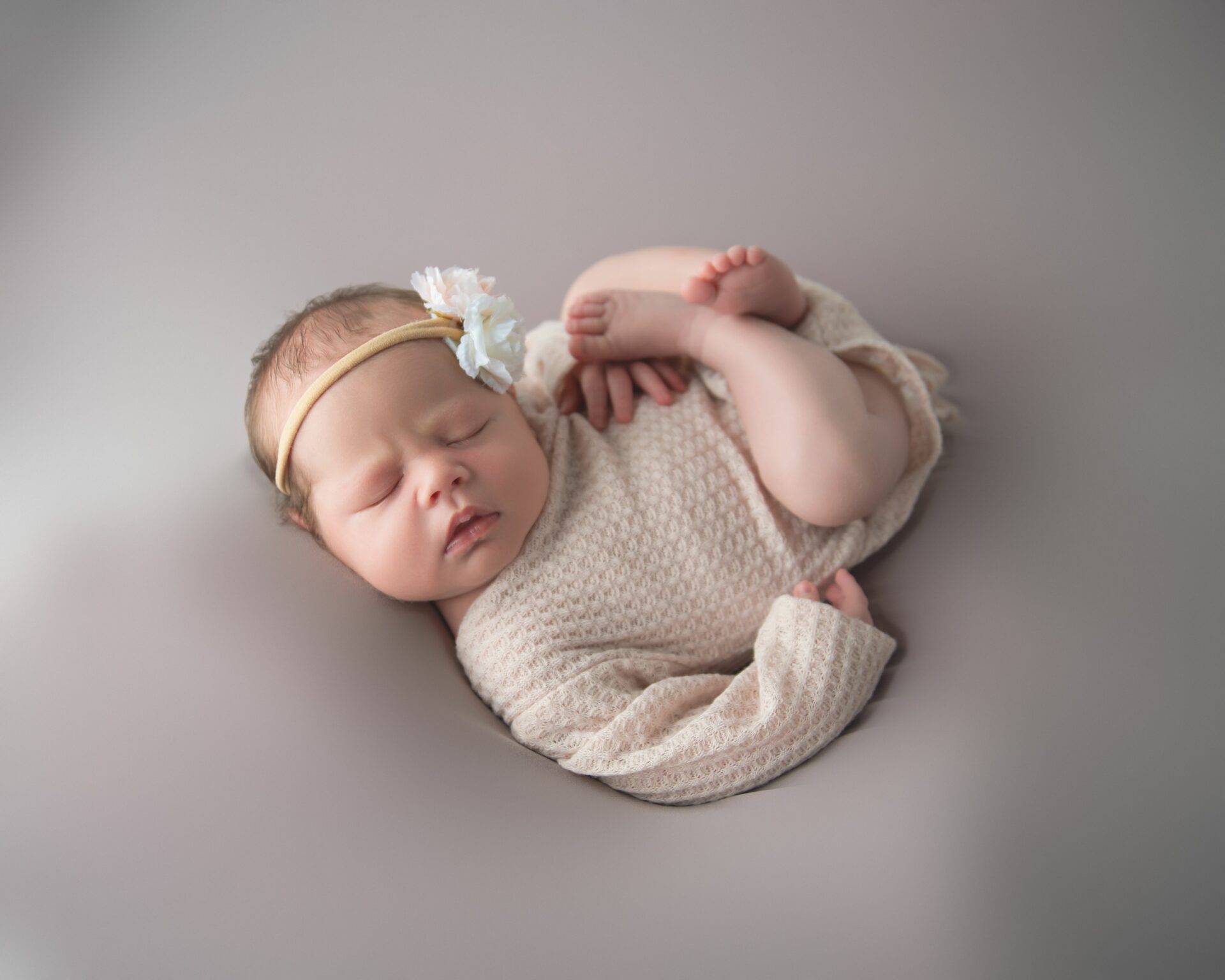 Sleeping newborn in a knitted outfit with a floral headband, lying on a soft, neutral-toned surface.