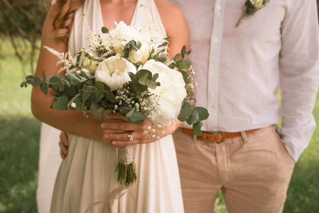 A couple stands together, the woman holding a bouquet of white flowers with greenery. They are dressed in light, casual attire, and are outside with grass in the background.