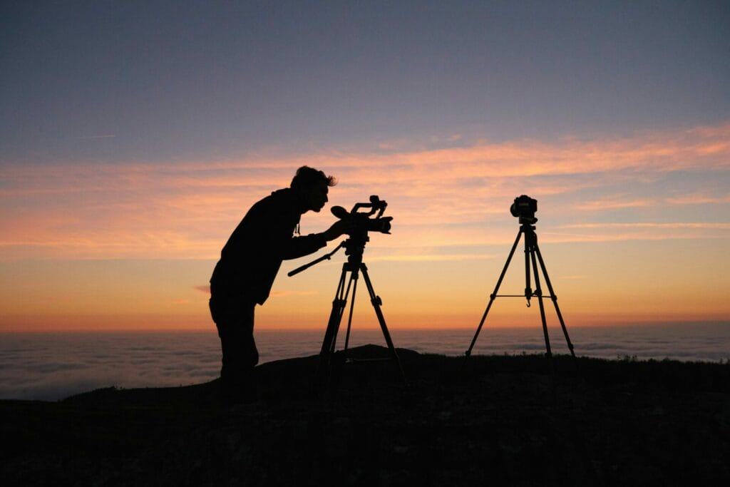 Silhouette of a person adjusting a camera on a tripod at sunset, with another tripod nearby against an orange and blue sky.