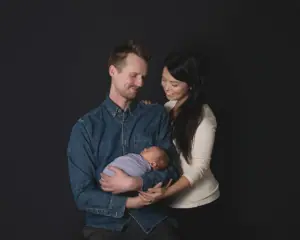 A seated man holds a sleeping baby in his arms, dressed in a blue onesie. A standing woman in a white top is beside him, leaning in with a smile. The background is black.