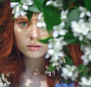 A person with red hair partially obscured by white flowers and leaves, looking directly at the camera.